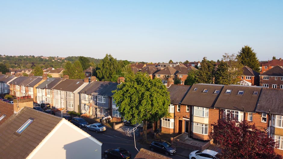 An aerial view of a residential street during daytime showing a row of terraced houses with brick and painted exteriors, pitched roofs with skylights, and large bay windows. Several parked cars line the narrow asphalt road in front of the homes. Prominently visible in the centre is a large, leafy green tree casting a shadow over the street, with additional trees and greenery dispersed throughout the neighborhood. The sky above is clear with a light blue hue, and the scene captures peaceful urban living. This image relates to house removals and home relocation services by illustrating the typical environment where furniture transport and packing logistics take place. Occasionally, Merton Man and Van provides home relocation and loading processes within such residential areas to ensure efficient moving, with a focus on careful handling of furniture and packing materials.'