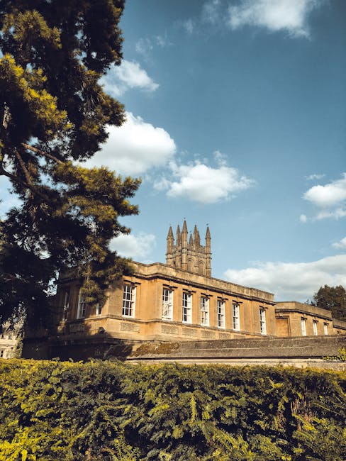 A historic stone building with large white-framed windows and a flat roof, partially obscured by dense green trees and shrubs in the foreground. The building features a prominent tower with gothic-sty
