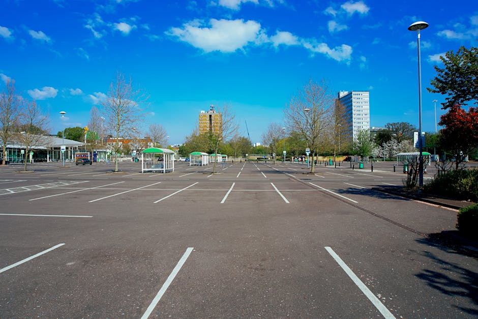 A large, mostly empty parking lot with clearly marked white parking spaces on asphalt ground, surrounded by a few leafless trees and some small structures with green roofs. In the background, there are multi-storey buildings under a bright blue sky with scattered white clouds. Tall streetlights are positioned along the edges of the lot, and some parked vehicles are visible towards the distance. The area appears well-maintained and spacious, providing ample room for home relocation activities such as loading and unloading furniture and packing supplies, which professional removals services like Merton Man and Van often handle within such environments.