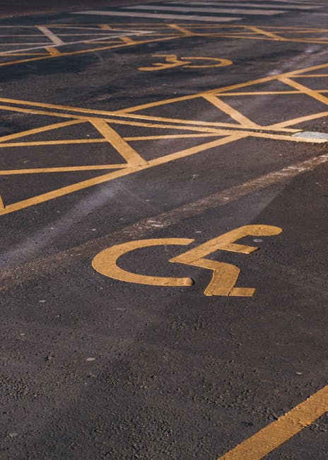 Close-up view of a black asphalt parking lot surface with multiple yellow painted parking space markings, including clearly visible disabled parking bay symbols featuring a wheelchair icon. The markings are arranged in a grid pattern with diagonal and horizontal lines, and some of the lines and symbols show slight wear and fading. The image captures the spatial arrangement of the parking bays, emphasizing the designated disabled parking space in the foreground. This setting likely represents an accessible parking area associated with residential or commercial premises where home relocation or furniture transport services by Merton Man and Van could occur, particularly during a home removal or moving process. The lighting appears natural with shadows indicating daylight conditions.