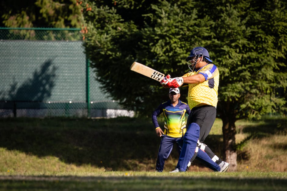 A cricket match is taking place on a green field with lush trees in the background. Two players are actively engaged in a game; the batter, dressed in white cricket whites, is in a batting stance, holding a cricket bat and preparing to hit a red cricket ball that is mid-air in front of him. The wicketkeeper, also in white clothing and wearing protective gloves and pads, is positioned behind the wickets, crouching slightly, ready to stop or catch the ball. The scene is well-lit with natural daylight, and the grass appears neatly maintained. Nearby, the boundary fence encloses the field, with parked cars and some spectators visible beyond the trees. The image captures a moment of dynamic action during an outdoor cricket match, illustrating the sport's equipment, players' focus, and the open, sporting environment, which could be relevant in content about sports facilities or recreational activities in relation to house removals or moving logistics.
