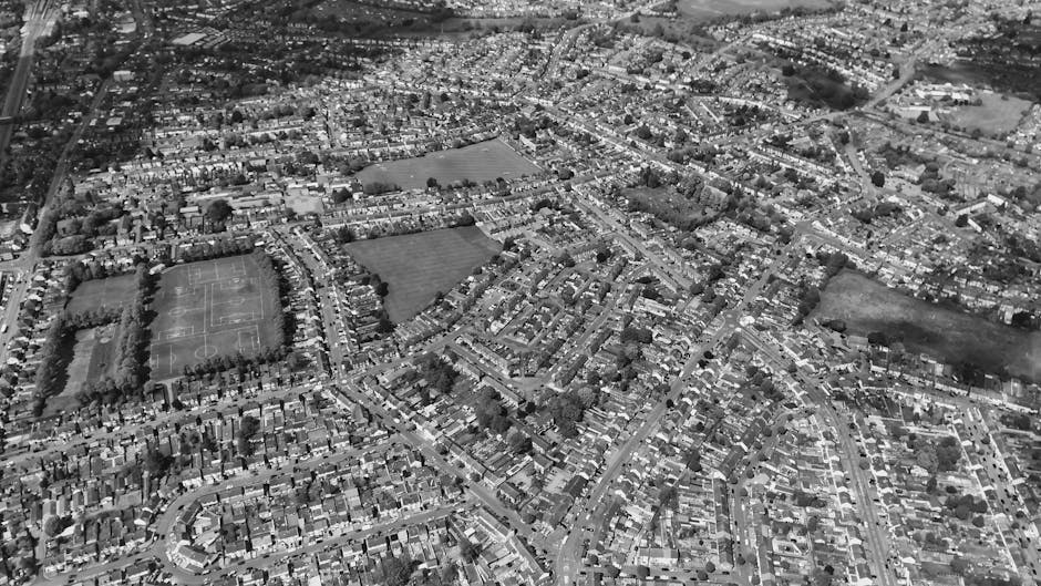 An aerial black-and-white photograph showing a residential area with densely packed houses arranged along winding streets, interspersed with green spaces and sports fields. The scene includes a large park with sports courts and open grassy areas, surrounded by an extensive network of roads and residential buildings. The image captures a suburban neighbourhood during daylight, with no visible vehicles or people. This view illustrates the typical layout of urban housing with tightly grouped dwellings, as seen in Morden SM4, relevant to house moves such as those offered by Merton Man and Van, who specialise in home relocation and furniture transport services.