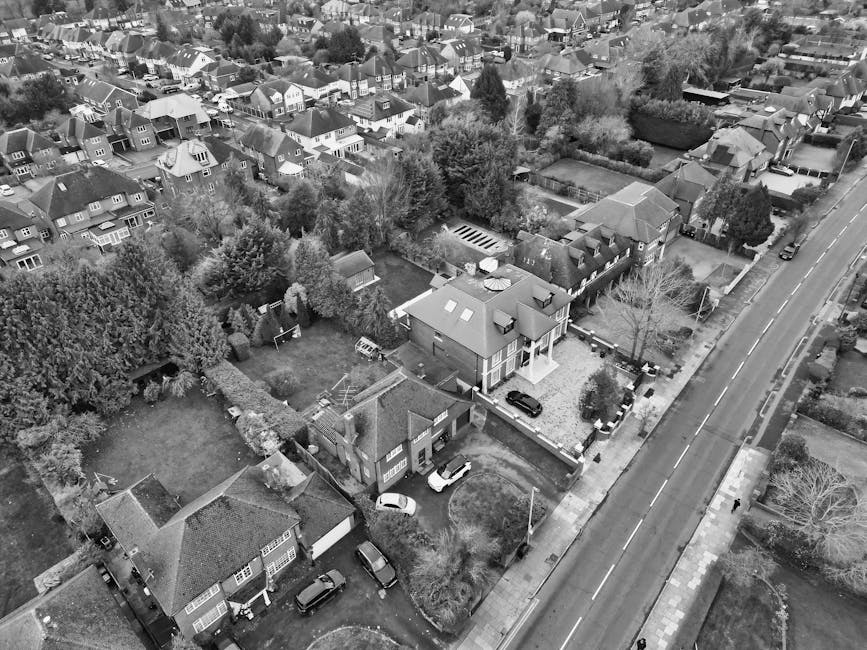 Aerial black-and-white photograph of a residential area showing multiple houses with pitched roofs, gardens, and trees. The central focus is on a two-storey house with a driveway, where several cars are parked. The house is surrounded by a fence, and there are visible lawn areas and small outbuildings in neighboring gardens. Adjacent to the property, a narrow street runs parallel, with additional parked vehicles along the pavement. The image captures the outdoor space, including garden fencing, trees, and paved pathways, illustrating a typical suburban setting suitable for house removals and home relocation services. The surroundings are calm, with natural light highlighting the layout of the neighborhood, supporting the context of furniture transport and packing and moving activities overseen by Merton Man and Van, as seen in the latest Morden SM4 moves from Morden Hall Park to home, Merton.