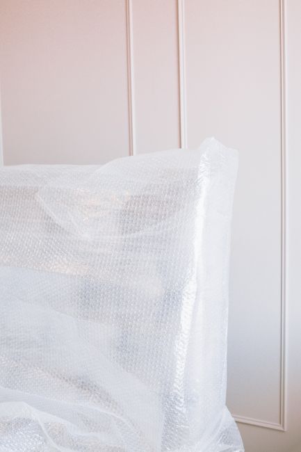 A large, wrapped antique wooden piano secured in protective plastic and bubble wrap, positioned inside a residential property near a white-paneled wall. The piano is being prepared for moving or transport, with part of it extending beyond the frame of the image. The wrapping materials indicate careful packing to prevent damage during the loading process. The background shows a clean, light-colored interior space, suggesting an organized house relocation setting. This image illustrates the detailed and fragile packing involved in furniture transport services, as provided by Merton Man and Van, specialized in house removals and furniture transport in Merton.