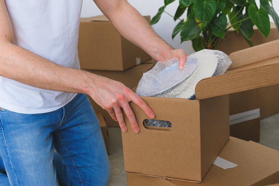 A person wearing a white t-shirt and blue jeans is seen during a home relocation process, carefully unwrapping a fragile item wrapped in bubble wrap from a cardboard box. The box is situated on a wooden table or furniture piece inside a room with a green leafy plant visible in the background. Additional cardboard boxes are present nearby, some closed and others open, indicating ongoing packing activities. The lighting in the space is natural, highlighting the careful handling of the delicate item, which might be a piece of fragile furniture or an antique. This scene reflects the meticulous packing procedures involved in furniture transport and moving services offered by Merton Man and Van, ensuring fragile items like antiques and pianos are prepared securely for transportation during a furniture removal or house move.