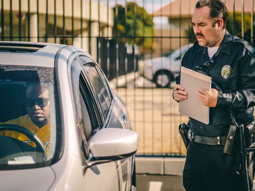 A police officer standing outdoors next to a parked silver vehicle during a home relocation scene, holding a clipboard and engaging with the driver who is seated inside the car. The driver, a person wearing sunglasses and a yellow shirt, is visible through the closed window. The scene takes place on a paved area with a metal fence and several other parked vehicles in the background. The police officer is dressed in a dark uniform with a badge and utility belt, while the vehicle is positioned close to the curb, indicating a loading or unloading process related to furniture transport or packing and moving activities. The lighting suggests daytime with natural sunlight, emphasizing the context of parking enforcement during removals, as highlighted by the webpage 'Parking enforcement during moves in Merton: Stay ticket-free, Merton' on mertonmanandvan.org.uk.