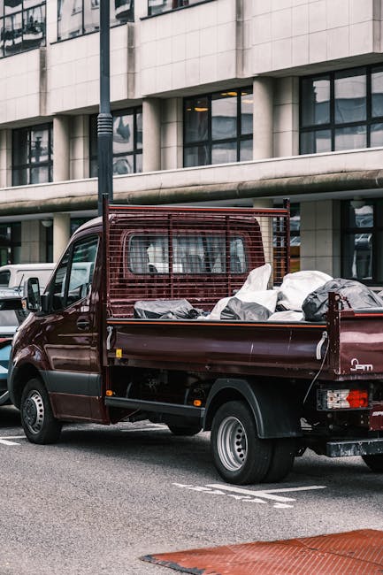 A dark red flatbed van parked on a city street during daylight hours, loaded with various packed items including large cardboard boxes, wrapped household belongings, and plastic bags, all covered with protective materials such as bubble wrap and blankets. The loading area of the van extends into the street, with the items arranged securely inside. In the background, modern office buildings with large glass windows and concrete facades can be seen, along with other vehicles parked nearby. The scene depicts the process of home removal or furniture transport, typical of house relocation services, with visible equipment like straps likely used for securing the load. This image is relevant to the services provided by Merton Man and Van, highlighting their capacity for efficient packing and loading during same-day emergency removals in Merton.