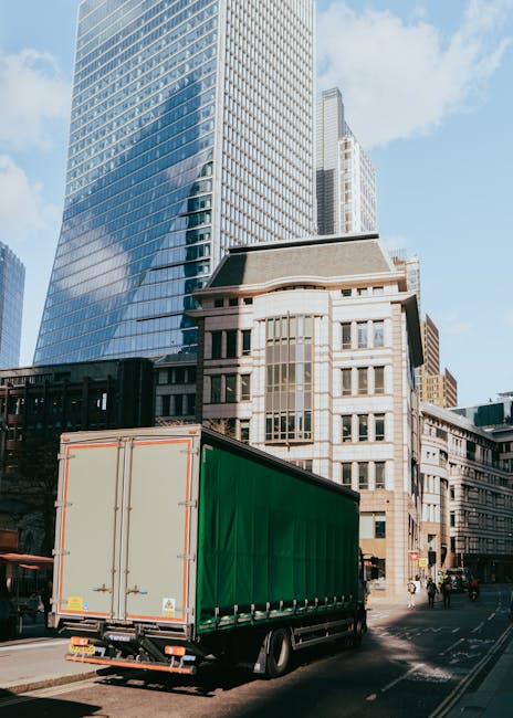 A large green and white delivery truck with closed rear doors is parked on a city street in front of modern high-rise buildings and older architectural structures. The truck is positioned on the right side of the image, with the rear facing the viewer and the front extending into the distance. The street is lined with pavement, and some pedestrians and cyclists are visible in the background. The surrounding scene features tall, glass-covered skyscrapers reflecting the blue sky and sunlight, alongside older, beige stone buildings. This setting illustrates urban delivery logistics, which may be part of a home removal or furniture transport process supported by Merton Man and Van. The scene emphasizes the loading and unloading environment during house moves within a city context, with the truck likely used for packing, moving boxes, furniture, and household items efficiently and securely during a relocation or same-day emergency removal.