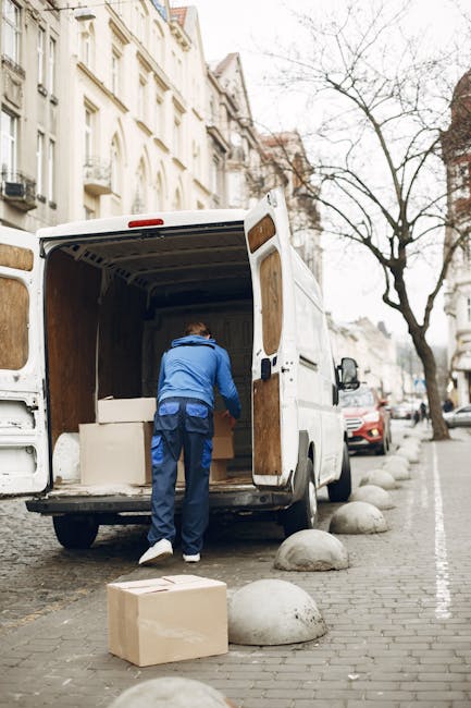 A dark red flatbed van parked on a city street during daylight hours, loaded with various packed items including large cardboard boxes, wrapped household belongings, and plastic bags, all covered with protective materials such as bubble wrap and blankets. The loading area of the van extends into the street, with the items arranged securely inside. In the background, modern office buildings with large glass windows and concrete facades can be seen, along with other vehicles parked nearby. The scene depicts the process of home removal or furniture transport, typical of house relocation services, with visible equipment like straps likely used for securing the load. This image is relevant to the services provided by Merton Man and Van, highlighting their capacity for efficient packing and loading during same-day emergency removals in Merton.