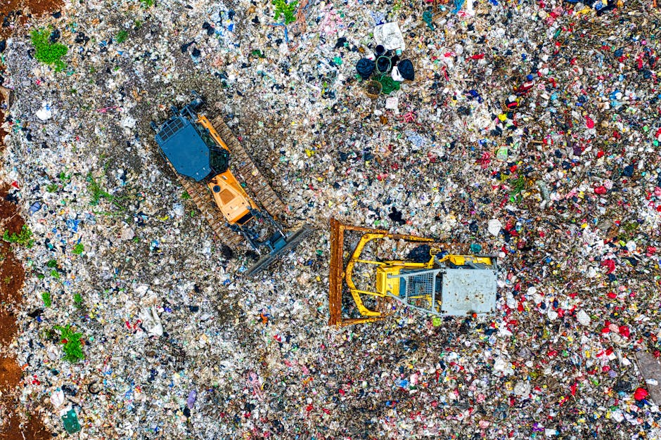 An aerial view of a large waste disposal site showing abundant mixed rubbish including plastic bottles, cardboard boxes, fabric scraps, and miscellaneous debris spread across the ground. Two heavy machinery vehicles—an orange tracked excavator and a yellow bulldozer—are actively engaged in managing the waste, with the excavator's arm extended and the bulldozer pushing or compacting materials. The scene is outdoors with natural lighting, situated on uneven, dirt-covered terrain. There are no buildings or structures visible, just the cluttered landscape of refuse. Occasionally visible are small patches of green vegetation emerging through the waste. The image illustrates the process of waste handling, which is relevant to the subject of waste disposal laws in Merton and proper waste management during house removals, as managed by [COMPANY_NAME], supporting proper relocation and packing practices.