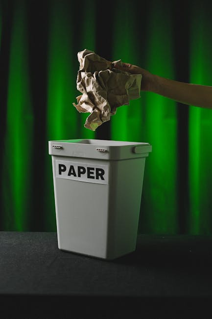 A hand holding a crumpled piece of brown paper is positioned above a white waste bin labeled 'PAPER' with black text. The bin is placed on a dark surface against a background of green curtains. The scene appears to be part of a home or office setting, illustrating the act of disposing of paper waste. This image reflects essential aspects of household or office waste management, which are relevant to house removals and moving services provided by Merton Man and Van. The focus on paper recycling highlights compliance with waste disposal laws in Merton to avoid fines after moving, aligning with the services related to packing and moving logistics.