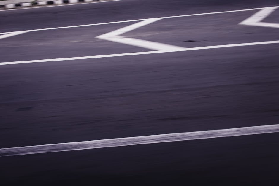 A blurred view of a dark asphalt street surface with white painted lane markings and arrows, indicating a turning lane or direction guidance. The pavement appears smooth with some visible textures, and the white lines contrast sharply against the dark road. The image captures motion or a lack of focus, with no vehicles or pedestrians visible. The scene is in an urban environment, possibly near residential or commercial buildings, related to house removals or relocation services, as seen in the website context of Merton Man and Van, specializing in furniture transport and home relocation logistics.