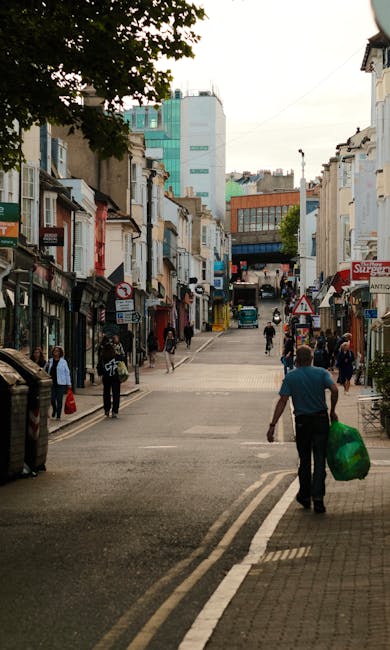 A busy narrow street in Wimbledon SW19, featuring a mix of small retail shops and residential buildings with various storefronts and signs. Pedestrians are walking on both sides of the street, some carrying shopping bags or personal belongings. The pavement is lined with parked bicycles and small outdoor displays. In the foreground, a man is walking away from the camera, carrying a large green bag, potentially during a home relocation or furniture transport process. Taller modern office buildings are visible further down the street, contrasting with the older, low-rise structures. Overhead, a cloudy sky provides diffuse lighting, and the scene depicts typical urban street activity related to house removals and local moving services, such as those offered by Merton Man and Van, highlighting the environment where furniture transport and packing may occur during house removals.